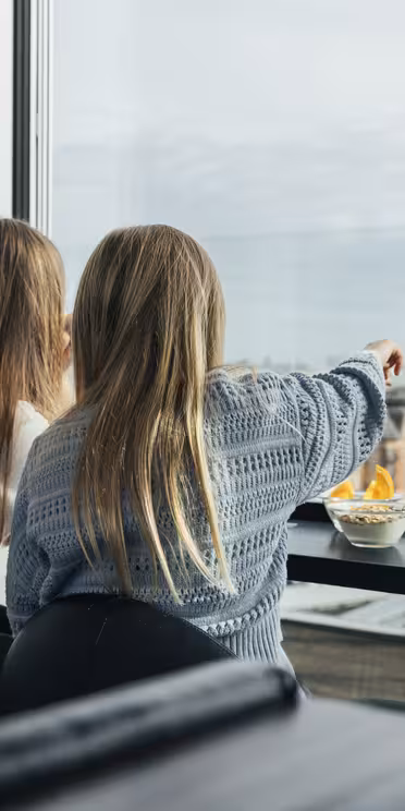 To personer med langt hår sitter ved et høyt bord ved et stort vindu med utsikt over et bybilde. Den ene peker ut mens den andre lytter. En skål med snacks står på bordet. Atmosfæren er avslappet og uformell.