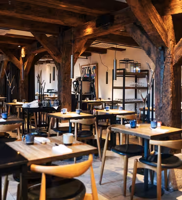 En rustikk, tom restaurant med trebord og trestoler. Synlige trebjelker og dempet belysning skaper en koselig atmosfære. Bord er dekket med blå kopper og sorte dekkebrikker, og hyller med glass er synlige i bakgrunnen.