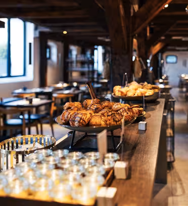 Et koselig bakeri med trebjelker og bord. En lang disk viser ulike bakverk, inkludert croissanter og danish. Klare glass og metallbeholdere er arrangert på siden. Myk belysning skaper en varm atmosfære.