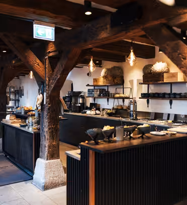 Et rustikk restaurantinteriør med synlige trebjelker, et bufféoppsett med ulike retter på mørke benker og pendelbelysning. Plassen har en koselig, varm atmosfære med hyller og dekorative elementer langs veggene.