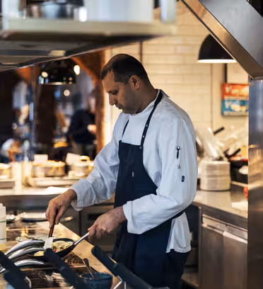 En kokk i hvit jakke og svart forkle lager mat på et restaurantkjøkken. Han er fokusert på å lage mat i en panne på komfyren. Kjøkkenet har overflater i rustfritt stål og hyller stablet med tallerkener og ingredienser.