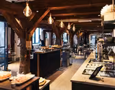 Romslig, rustikk restaurantinteriør med trebjelker og industriell belysning. Et bufféoppsett med ulike retter vises på diskene. Bord og stoler er arrangert i hele rommet med store vinduer som gir naturlig lys.