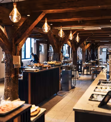 Romslig, rustikk restaurantinteriør med trebjelker og industriell belysning. Et bufféoppsett med ulike retter vises på diskene. Bord og stoler er arrangert i hele rommet med store vinduer som gir naturlig lys.