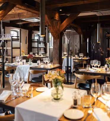 Koselig restaurantinteriør med trebjelker og varm belysning. Bordene er dekket med hvite duker, vinglass og små blomsterdekor. Atmosfæren er intim og innbydende.