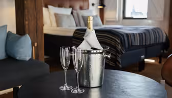 En flaske champagne pakket inn i en serviett står i en isbøtte på et rundt bord, med to tomme champagnefløyter ved siden av. Bakgrunnen viser et koselig soverom med seng, stripete sengetøy og puter, opplyst av naturlig lys fra et vindu.