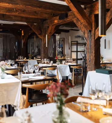 Et koselig restaurantinteriør med trebjelker og flere bord dekket med hvite duker, glass og blomsterdekor. Myk belysning skaper en varm atmosfære, og hyller med flasker i bakgrunnen øker stemningen.