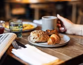 Tallerken med en croissant, rugbolle og wienerbrød på et trebord, sammen med en avis, bestikk og et glass. En person holder et grått krus. Uskarp bakgrunn med varm belysning.