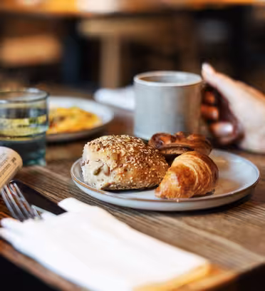 Tallerken med en croissant, rugbolle og wienerbrød på et trebord, sammen med en avis, bestikk og et glass. En person holder et grått krus. Uskarp bakgrunn med varm belysning.