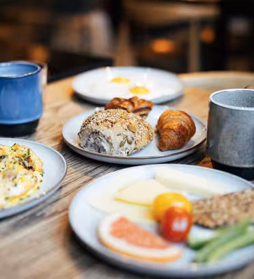 Et bord med frokostartikler, inkludert en omelett, diverse brød, ost, delikatessekjøtt, grønnsaker og drikke i krus og glass. Innstillingen er innendørs med en varm, innbydende atmosfære.
