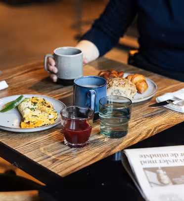 En person i en mørk genser sitter ved et trebord med et måltid. Bordet har en omelett, croissanter, kaffe, juice og vann. En avis er delvis synlig i forgrunnen. Servietter og gafler legges til siden.