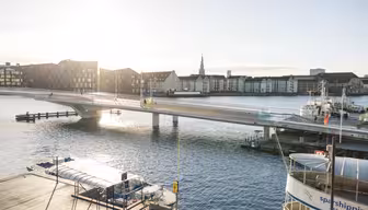 En solbelyst scene ved vannkanten med en moderne gangbro som strekker seg over en rolig elv. Bygninger langs vannet og et par båter ligger til kai i nærheten. Himmelen er klar og sola kaster refleksjoner på vannet.