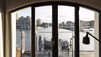 Utsikt gjennom et buet vindu som viser en scene ved vannet med moderne og tradisjonelle bygninger. En rolig elv reflekterer den overskyede himmelen, mens en strandpromenade med mennesker gir liv til det rolige bylandskapet.
