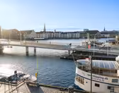Utsikt over en rolig havn med en bro som krysser vannet. Flere personer sykler og går på broen. Båter ligger til kai i nærheten, og historiske bygninger ligger langs den motsatte kysten under en klarblå himmel.