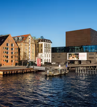 Vannfrontutsikt over moderne og historiske bygninger i København. Til venstre, fargerike tradisjonelle bygninger og båter. Til høyre strekker det slanke, glassveggede Kongelige Danske Skuespilhus seg over vannet under en klarblå himmel.