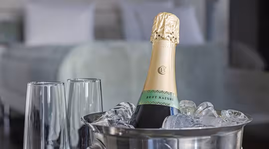 En flaske Brut Nature-champagne står avkjølt i en isbøtte på et bord. To tomme champagnefløyter står ved siden av. En mykt uskarp, veloppredd seng er synlig i bakgrunnen, noe som antyder en luksuriøs setting.