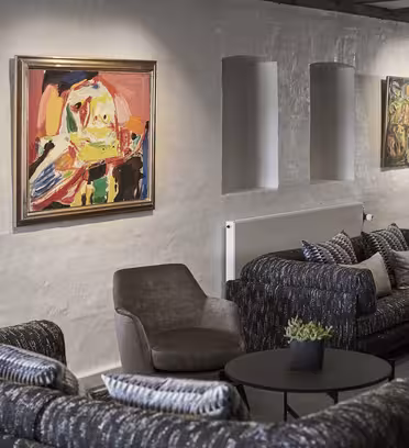 En moderne stue med abstrakte malerier på hvite vegger. To mørke sofaer og en grå lenestol omkranser runde sorte bord. Minimalistiske lamper og små potteplanter fullfører de koselige omgivelsene.