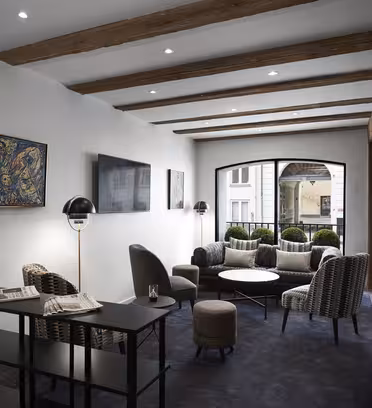 En moderne salong med synlige trebjelker, grått teppe og stilige møbler, inkludert lenestoler og et lite bord. Kunst pryder veggene, og naturlig lys strømmer inn gjennom store buede vinduer, og forsterker den koselige atmosfæren.
