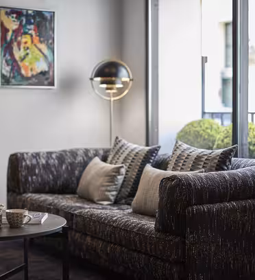 Stilig stue med mønstret sofa, geometriske puter og et rundt salongbord med tekanne og kopp. En moderne lampe står i nærheten. Et fargerikt abstrakt maleri henger på en blek vegg. Glassdører avslører en terrasse med pent trimmede busker.