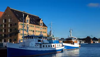 To blå og hvite båter kalt "BJØRNHOLM" ligger til kai ved siden av en pittoresk, historisk murbygning med topptak og flere vinduer. Scenen er satt under en klar blå himmel, og vannet reflekterer det sterke lyset.