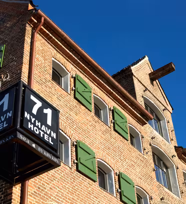 Murbygning med grønne skodder under en klar blå himmel. På et svart skilt med hvit tekst står det «71 Nyhavn Hotel». Bygget har buede vinduer og synlige trebjelker.