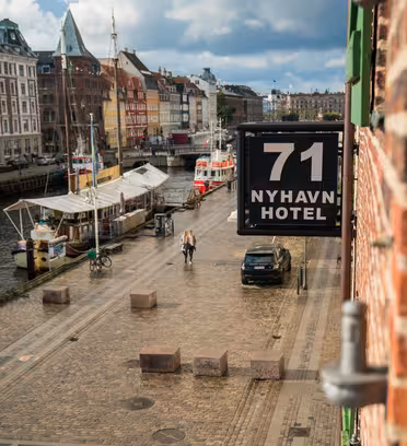 Utsikt over brosteinsgate langs en kanal med fortøyde båter i Nyhavn, København. Folk går langs stien, og fargerike bygninger langs vannkanten. På et skilt på murbygningen står det «71 Nyhavn Hotel». Skyet himmel i bakgrunnen.