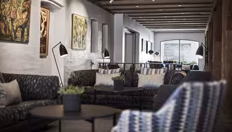 Elegant lobbyområde med moderne sofaer, runde svarte salongbord med potteplanter og fargerik abstrakt kunst på veggene. Synlige takbjelker og et stort buet vindu i bakgrunnen gir en koselig atmosfære.
