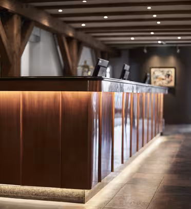 Et svakt opplyst hotellresepsjonsområde med en trepult opplyst nedenfra. To dataskjermer sitter på skrivebordet. Rommet har trebjelker, flislagte gulv og innrammede kunstverk på veggene.