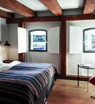 Et koselig hotellrom med en seng dekket av et stripete teppe, to puter og en tuftet hodegjerde. Trebjelker er synlige i taket. To små, buede vinduer med åpne skodder gir utsikt over et bybilde. Et skrivebord og en stol er i hjørnet.
