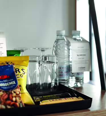 Et velkomstbrett på et hotellrom på et trebord med en flaske rødvin, to flasker vann, snackspakker, to tomme glass og en lapp til gjesten. Naturlig lys kommer fra et vindu i bakgrunnen.