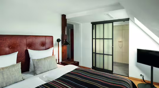 Et minimalistisk hotellrom med dobbeltseng med hodegjerde i brunt skinn og stripete sengetøy. Det er en skyvedør i glass som fører til et bad, en liten TV på stativ og en tresøyle nær sengen.