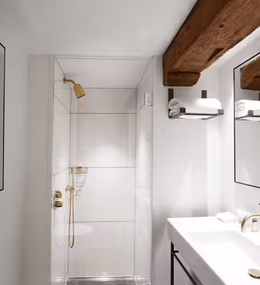 Et moderne bad med glassdusj med gullarmaturer, hvit vask med gullkran og to speil. Synlige trebjelker i taket gir et rustikk preg. Hvite vegger og fliser skaper et rent, minimalistisk utseende.