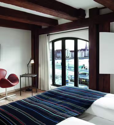 Hotellrom med trebjelker med en seng dekket av et stripete teppe, to brune stoler, et lite skrivebord med en lampe og en veggmontert TV. Store vinduer gir utsikt over en vannkant og bygninger utenfor.