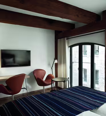 Et moderne hotellrom med synlige trebjelker, en seng med et stripete teppe, to oransje stoler, en veggmontert TV, et lite skrivebord og en gulvlampe. Høye vinduer viser utsikt over en nabobygning.