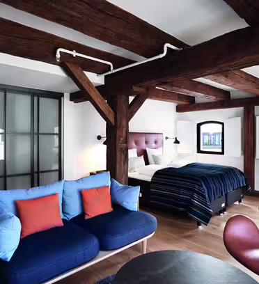Et koselig, rustikk soverom med trebjelker og dobbeltseng. Rommet har en blå sofa med oransje puter, en moderne stol og et lite rundt bord. Det er tre buede vinduer og bad med skyvedør.