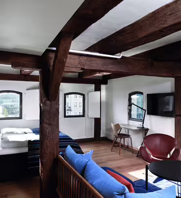 Et koselig, rustikk hotellrom med trebjelker, en seng med hvitt og blått sengetøy, en sofa, et lite rundt bord, stoler og en TV montert på veggen. Store vinduer gir utsikt over et urbant landskap utenfor.