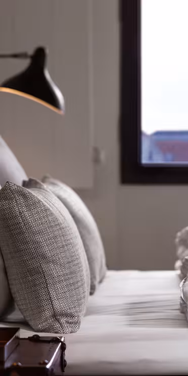 Et koselig soverom med en stor seng med hvitt sengetøy og grå puter. To moderne svarte vegglamper lyser opp rommet. Et vindu i bakgrunnen viser silhuetten av en fjern bygning mot en klar himmel.