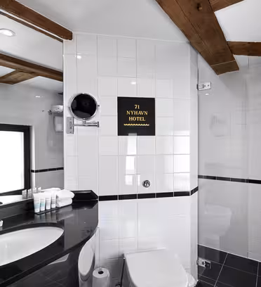 Et moderne bad med hvite flislagte vegger og trebjelker. Har en svart benkeplate med vask, badeprodukter og speil. Et lite dusjområde er synlig. På en tavle står det "71 Nyhavn Hotel." Håndklær er pent ordnet på et stativ.