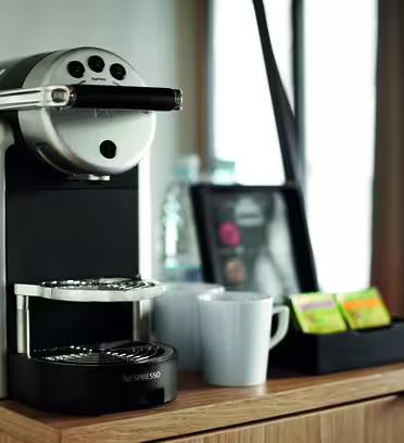 En kompakt espressomaskin står på en trebenk, ledsaget av to hvite krus og et brett med tepakker. En vannflaske og bakgrunnsbelyst vindu bidrar til den koselige atmosfæren.