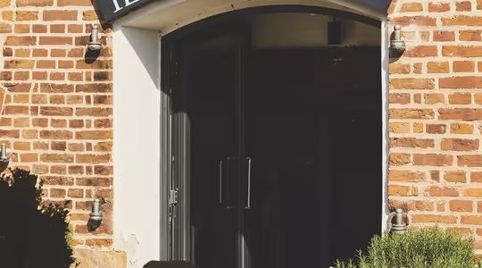 Murstein eksteriør av en restaurant med et svart skilt som leser "Ristorante Il Rosmarino" over inngangen. Uteservering inkluderer trebord satt med glass og servietter, under en stor paraply. Grønne omgivelser omgir området, og skaper en koselig atmosfære.