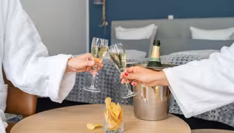To personer i hvite kapper klirrer i champagneglass på et hotellrom. En flaske champagne står i en isbøtte på et bord med et glass potetgull i forgrunnen. Sengen i bakgrunnen er pent laget.